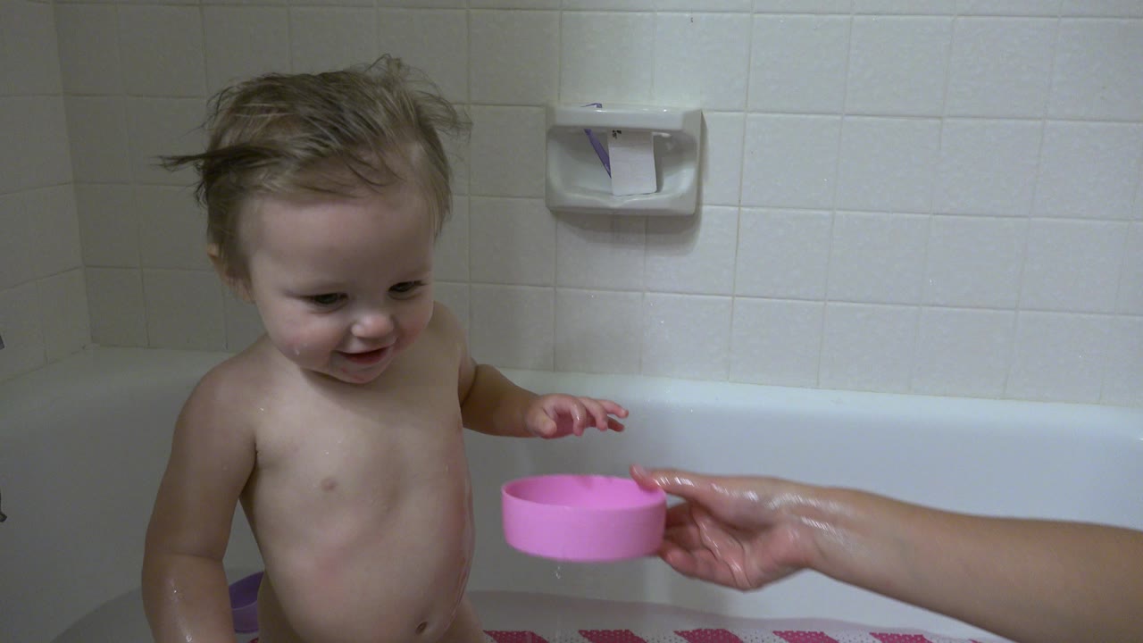 In Westminster, Colorado, the child smiles in the bathtub as an adult hand keeps a pink bowl close to the water against a white tile wall.