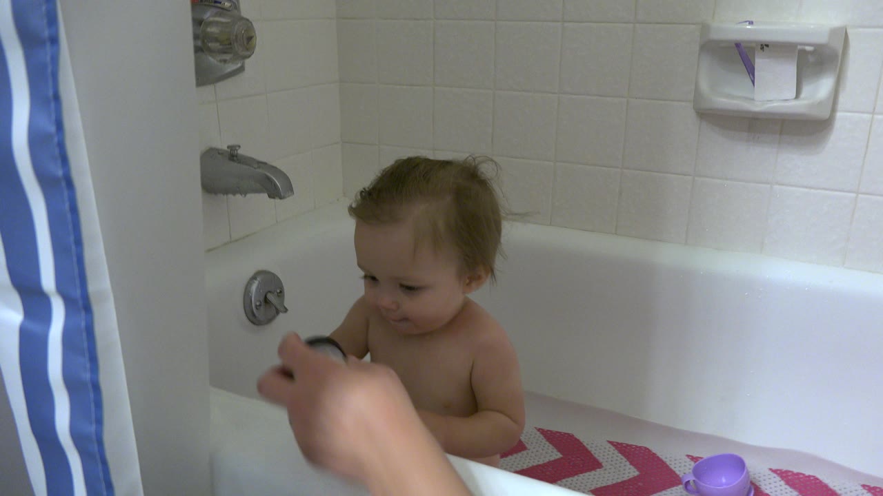 Baby sitting in a bathtub while an adult hand holds a bath toy nearby.