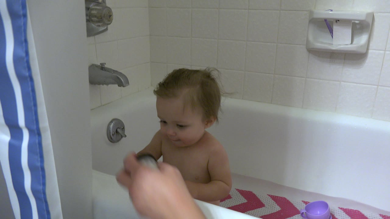 In Westminster, Colorado, the baby looks down toward an adult hand and toy while seated against white tile and a partly visible blue striped shower curtain.