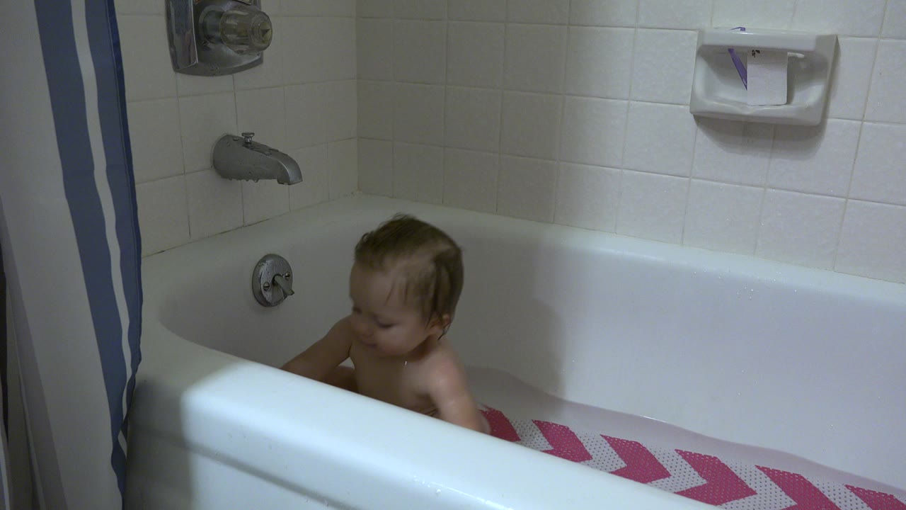 Baby seated low in a white bathtub beside a pink patterned towel.