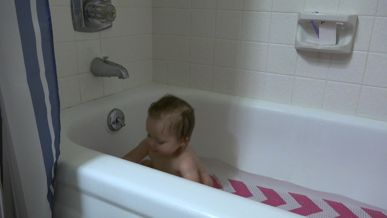 The baby remains seated in the Westminster bathroom bathtub, looking down as the tub rim, faucet hardware, white tile, and wall soap dish stay in view.