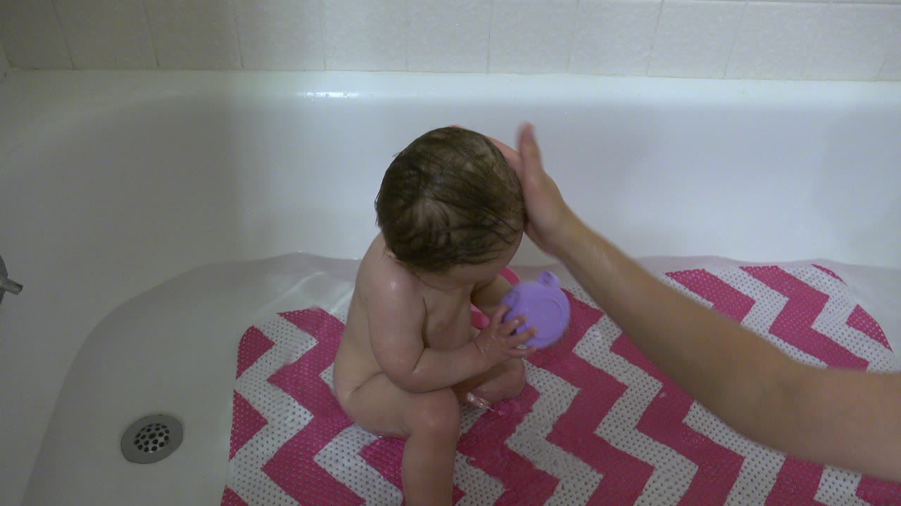 Baby in bathtub holding a purple cup as an adult hand reaches in.