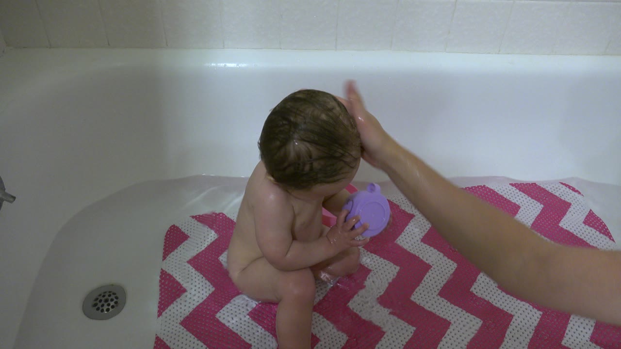 In Westminster, Colorado, the baby remains seated in the tub with the purple cup against the body as an adult hand touches the top of the head.