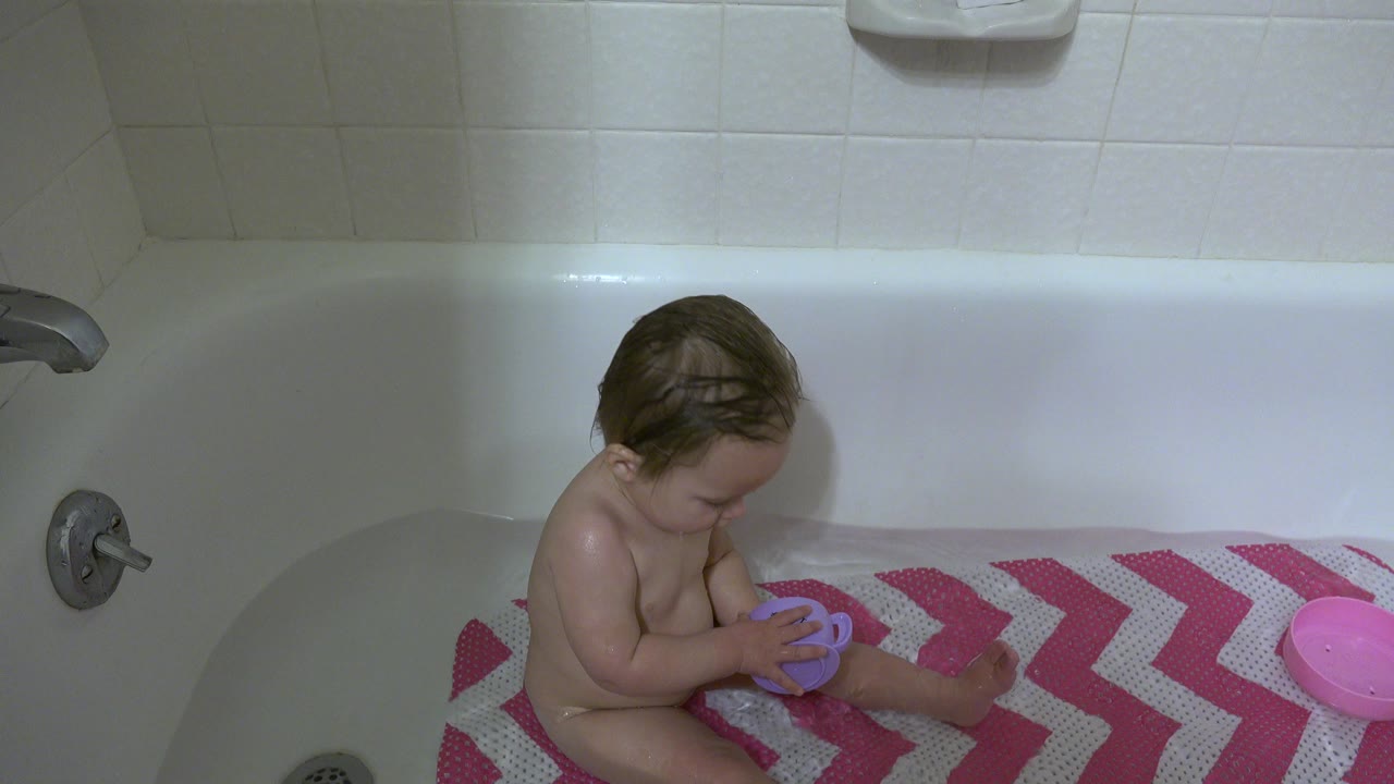 A baby sits low in a white bathtub in Westminster, Colorado, looking down while holding a purple mug on a pink-and-white chevron bath mat.