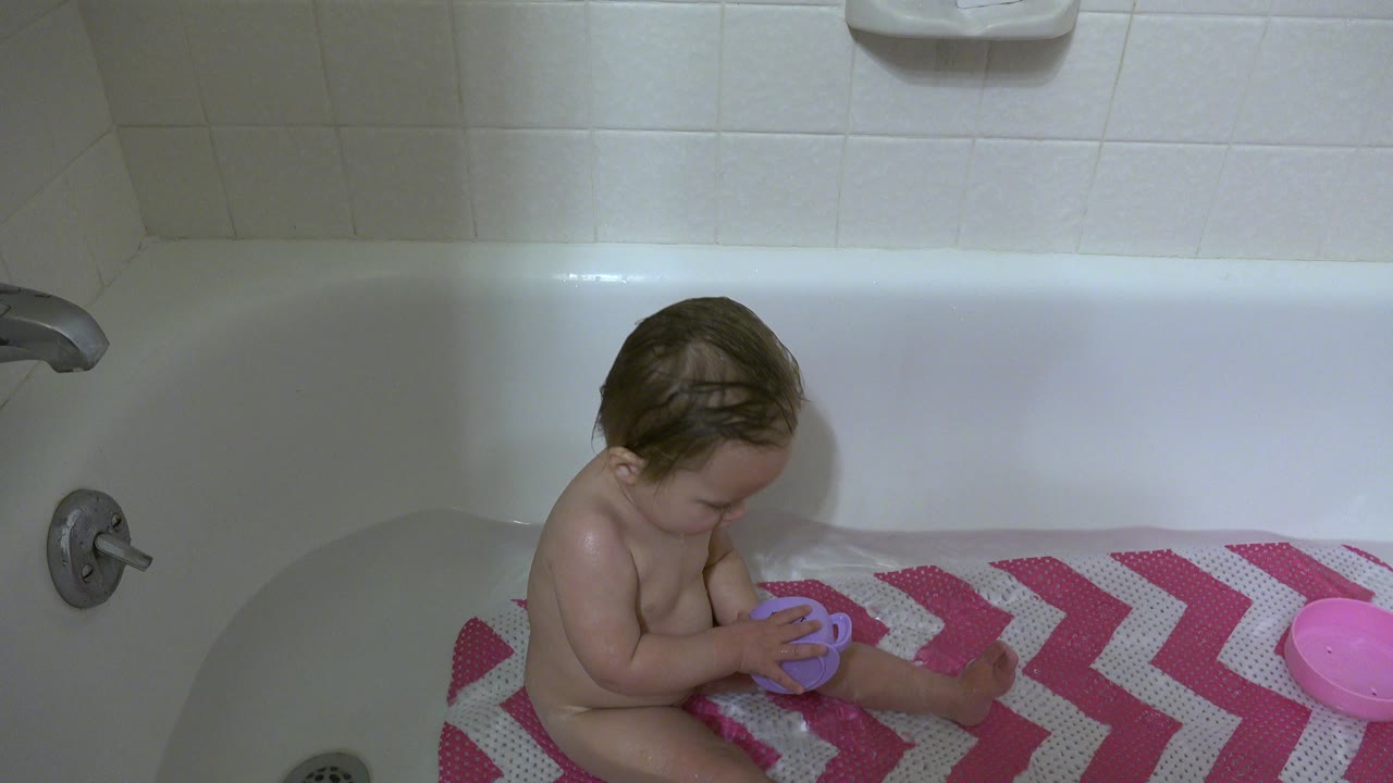 The baby remains seated in the Westminster bathtub, head tilted down toward the purple mug, with the faucet and pink chevron mat framing the bath scene.
