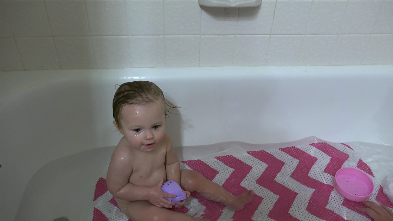 Toddler in a bathtub holding a purple cup beside a pink bowl.