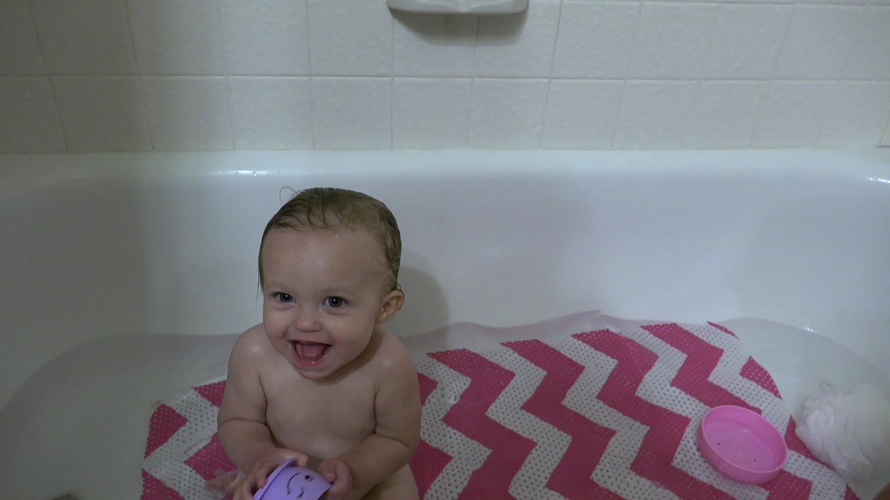 Smiling baby sitting in a bathtub with a purple cup.