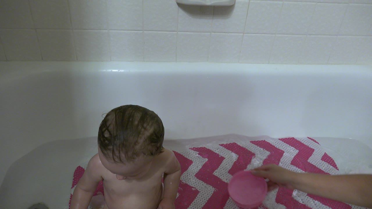 Baby sitting in a bathtub beside a pink-and-white chevron mat.