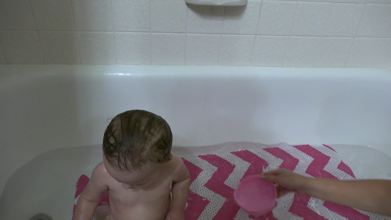 An adult arm reaches into the white bathtub with a pink cup while the baby sits near the left side of the frame in the Westminster, Colorado home bathroom.
