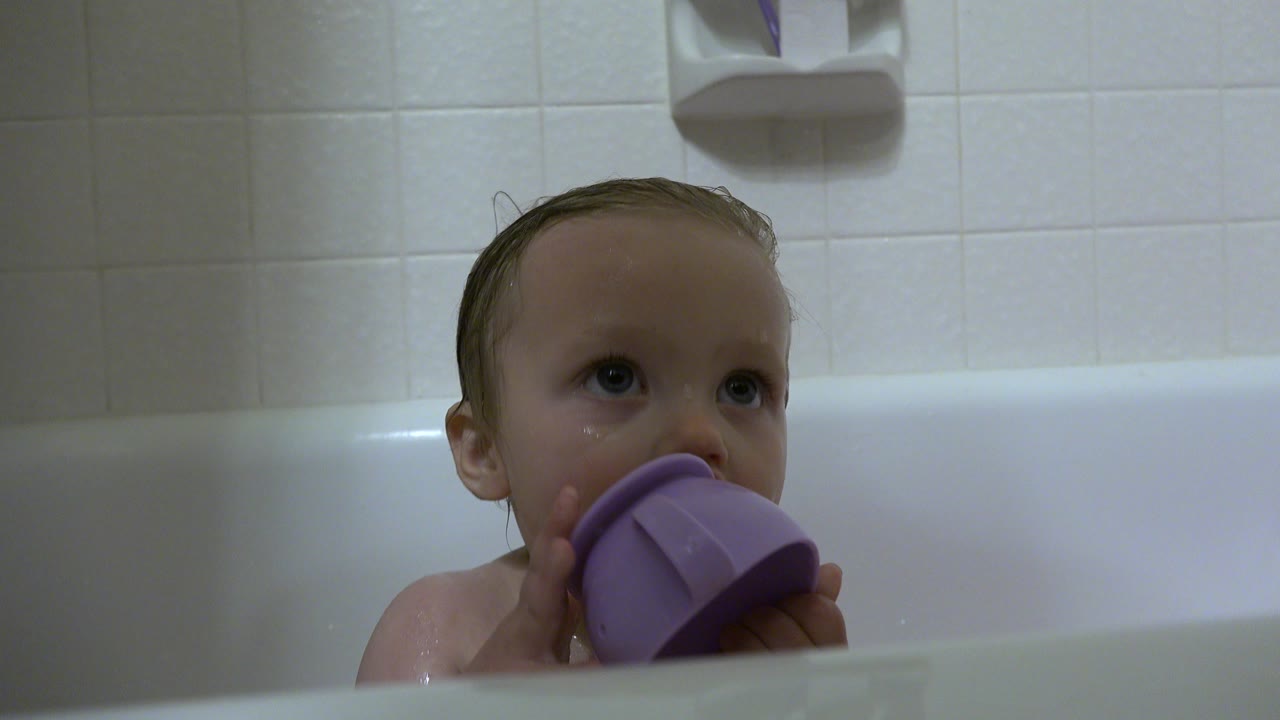 A wet baby boy sits low in a white bathtub in Westminster, Colorado, holding a purple cup near his mouth beneath pale bathroom tiles.
