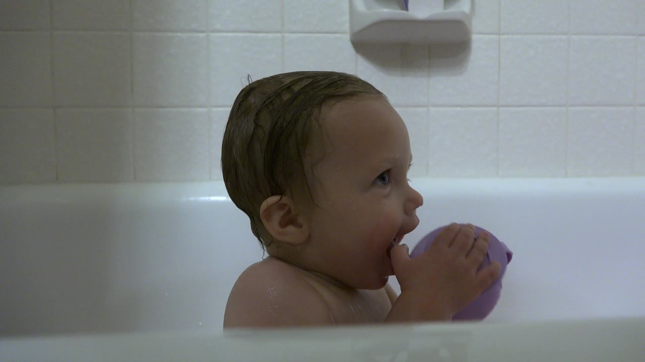Wet-haired baby sits in a white bathtub in Westminster, Colorado, holding a purple cup close to the mouth against a tiled bathroom wall.
