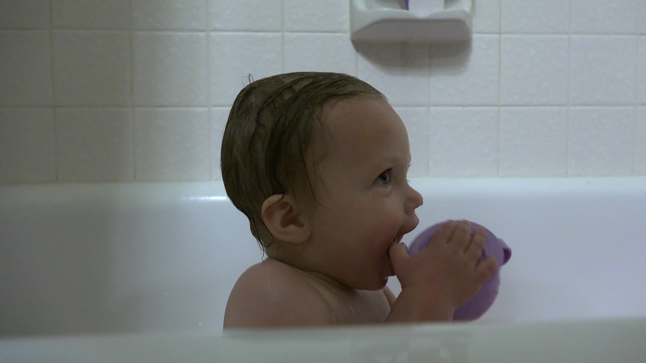 Baby remains in profile in the Westminster bathroom tub, with the purple cup lifted to the mouth and pale square tile behind.