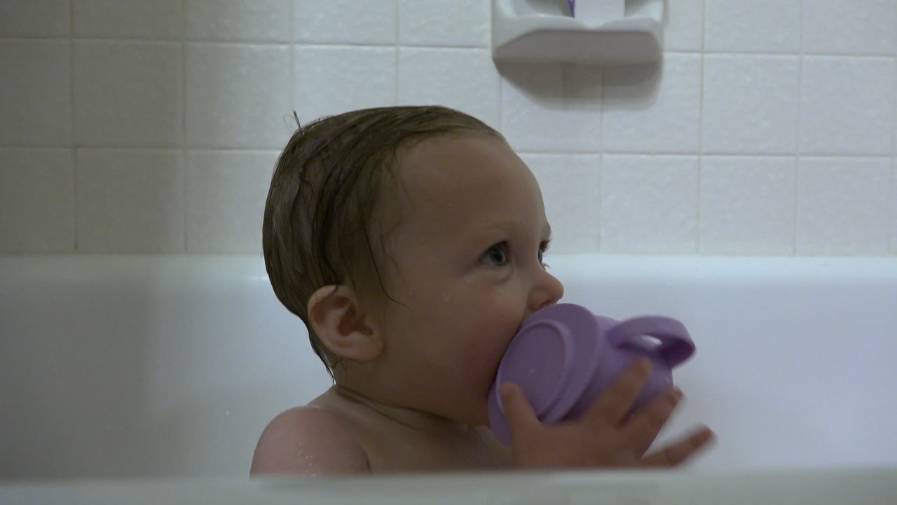 Baby in the white tub raises the purple handled cup higher to the mouth while looking right, with the tiled Westminster bathroom wall in the background.