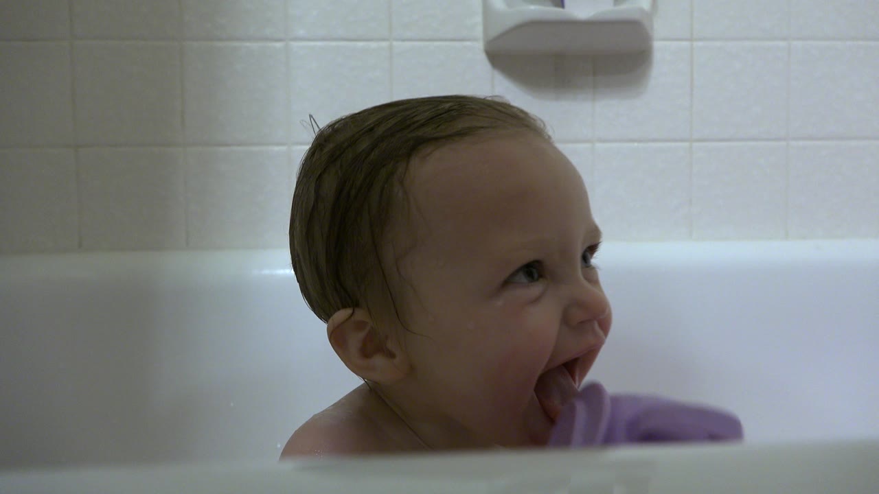 Baby opens mouth wide in a bathtub with purple cup partly visible.