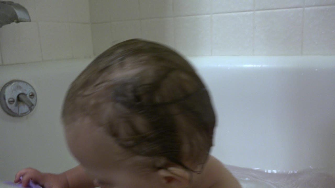Wet-haired baby leaning down in a white bathtub.