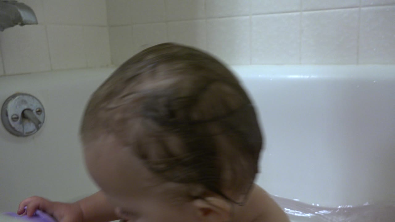The baby remains bent forward in the white bathtub in Westminster, Colorado, with a purple bath toy partly visible near the right edge.