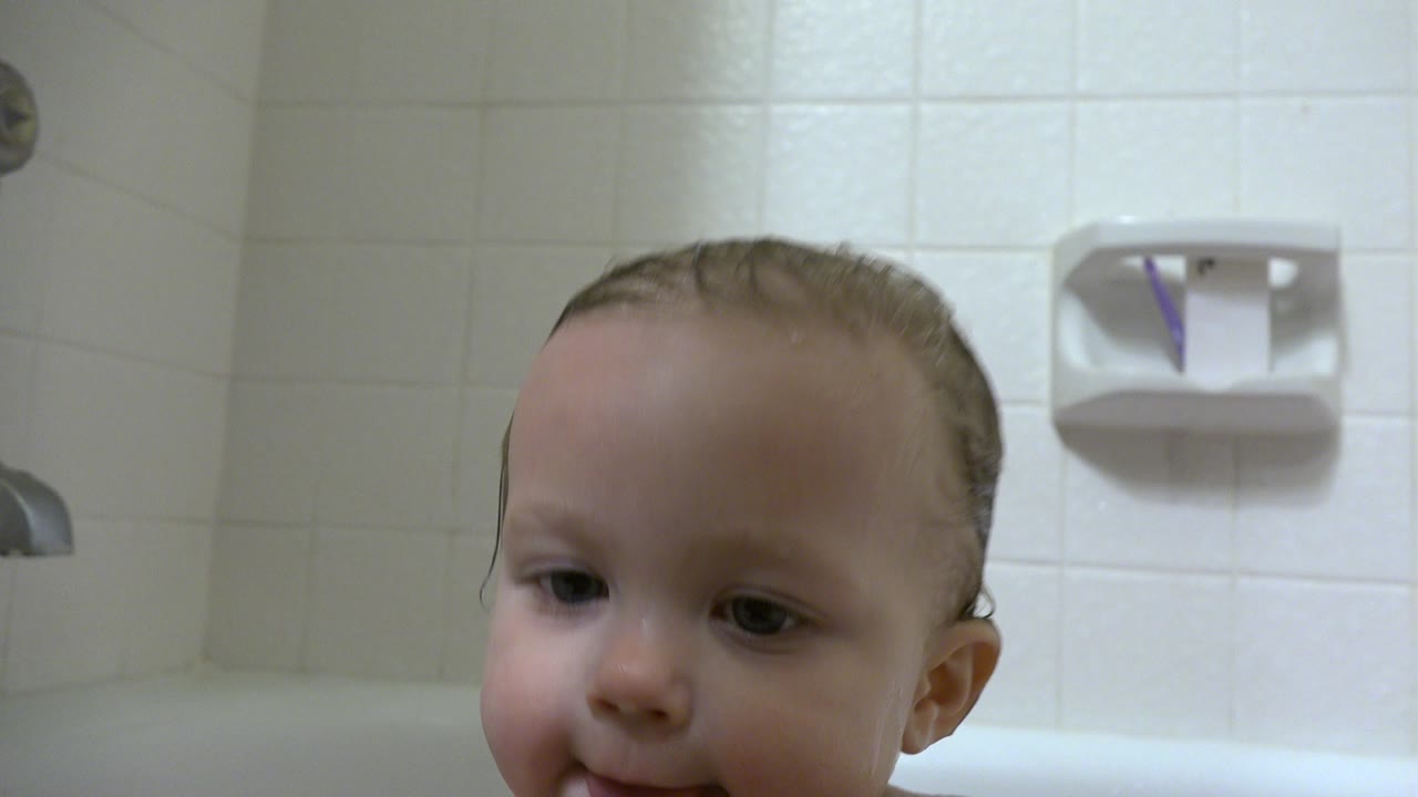 At 0:01, the baby's wet face and shoulders sit just above the bathtub edge, with pale tile and a wall soap dish behind.