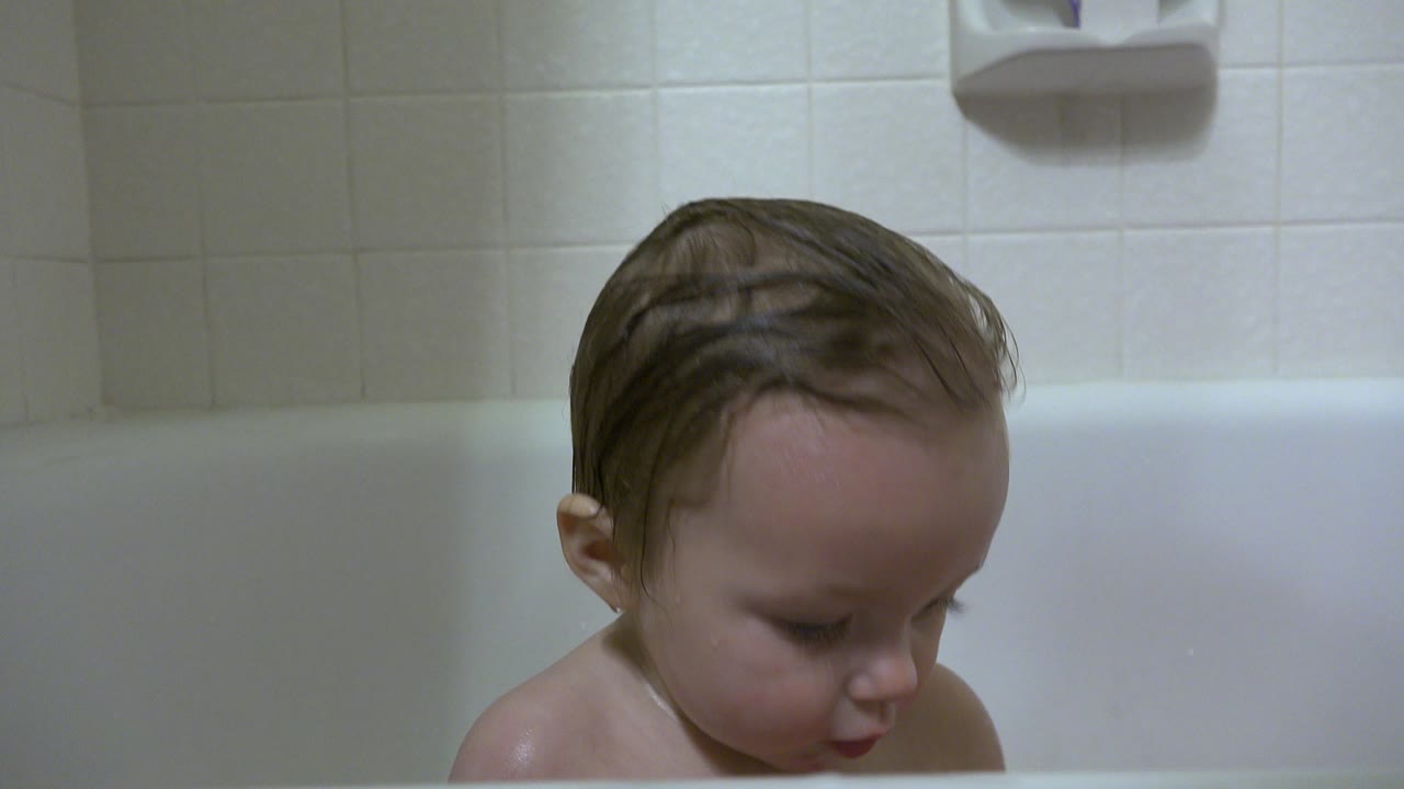 Wet-haired baby girl looking down in a white bathtub.