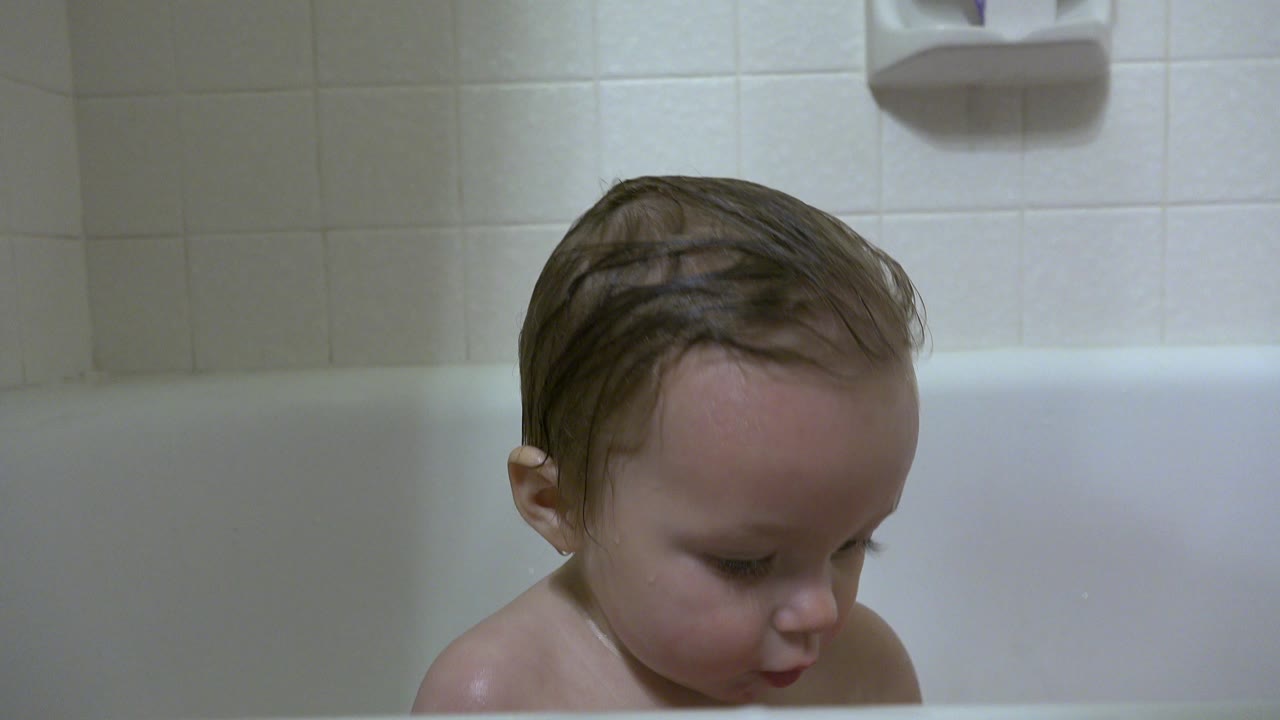 Baby girl with wet hair sits low in the tub in Westminster, Colorado, framed close against square bathroom tiles and soft overhead light.