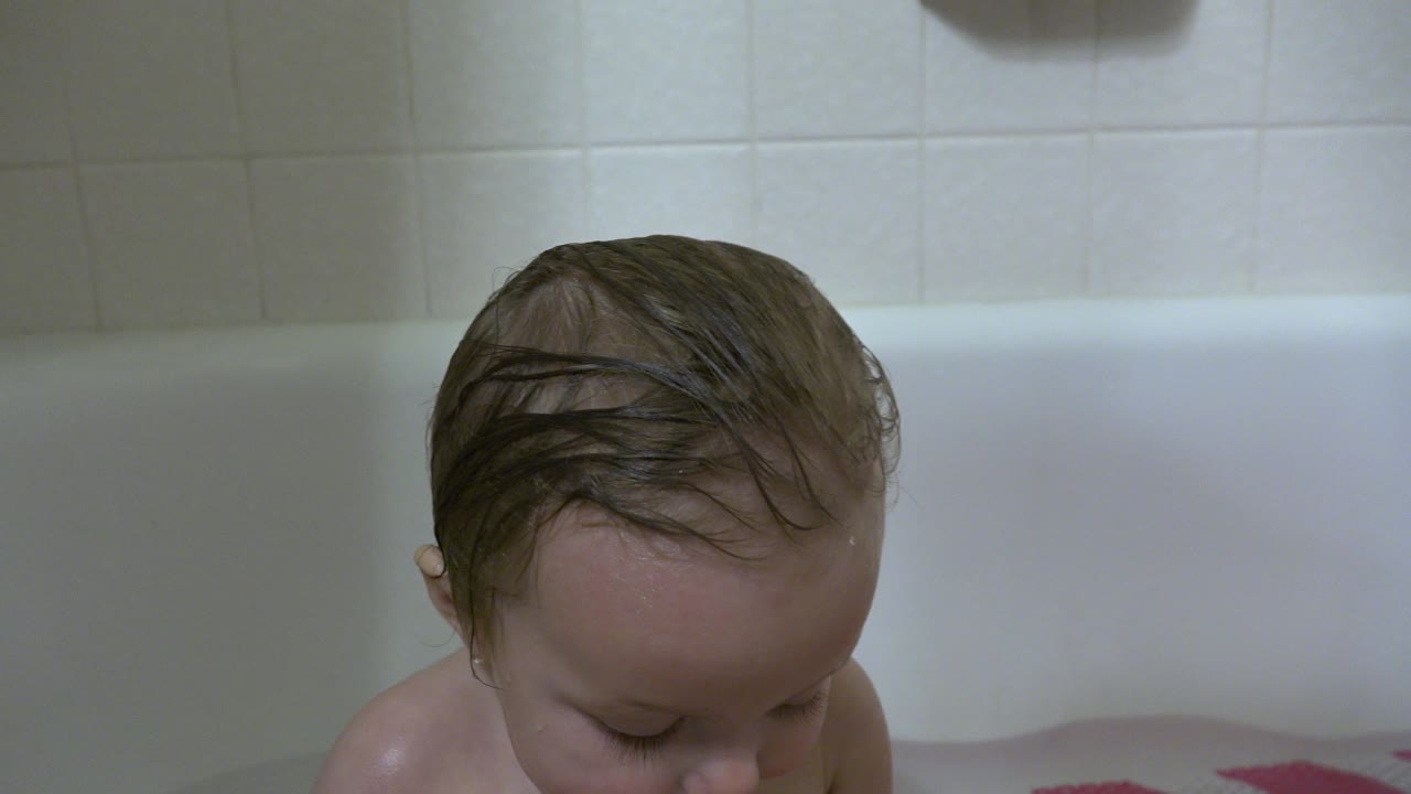 At 0:01, the baby remains low in the white bathtub in Westminster, Colorado, with wet hair combed across the head and the tiled wall softly lit behind.
