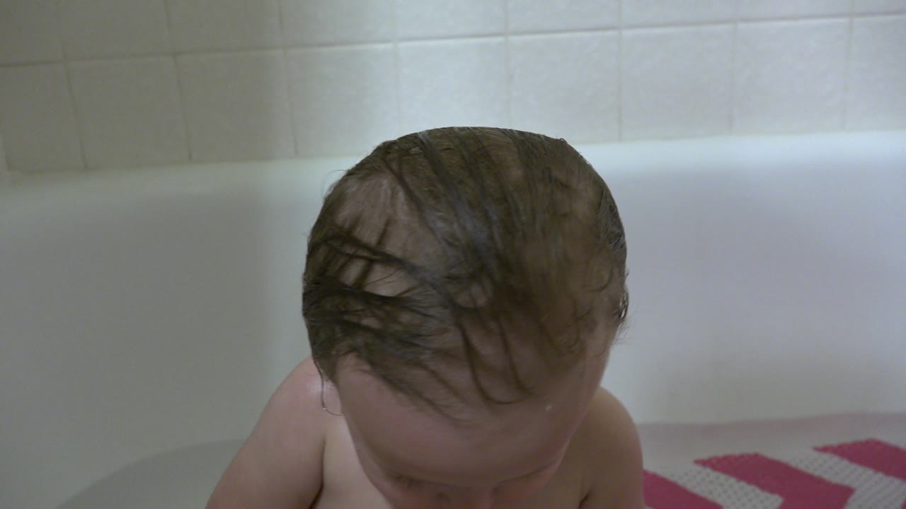 At 0:02, the baby tilts down in the white bathtub in Westminster, Colorado, with the crown of the wet head prominent and a pink bath mat partly visible at the side.