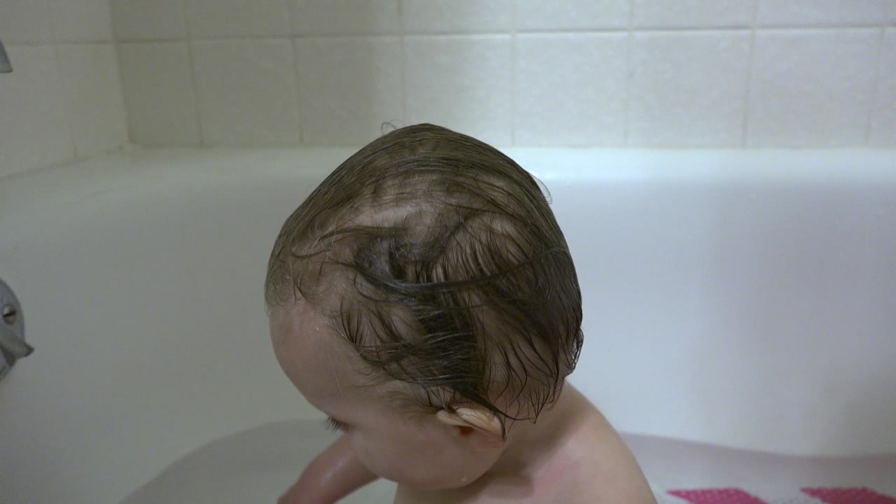 A close tub-level view shows the baby turned downward in the white bathtub, with wet hair, pale wall tile, and chrome hardware visible in Westminster, Colorado.