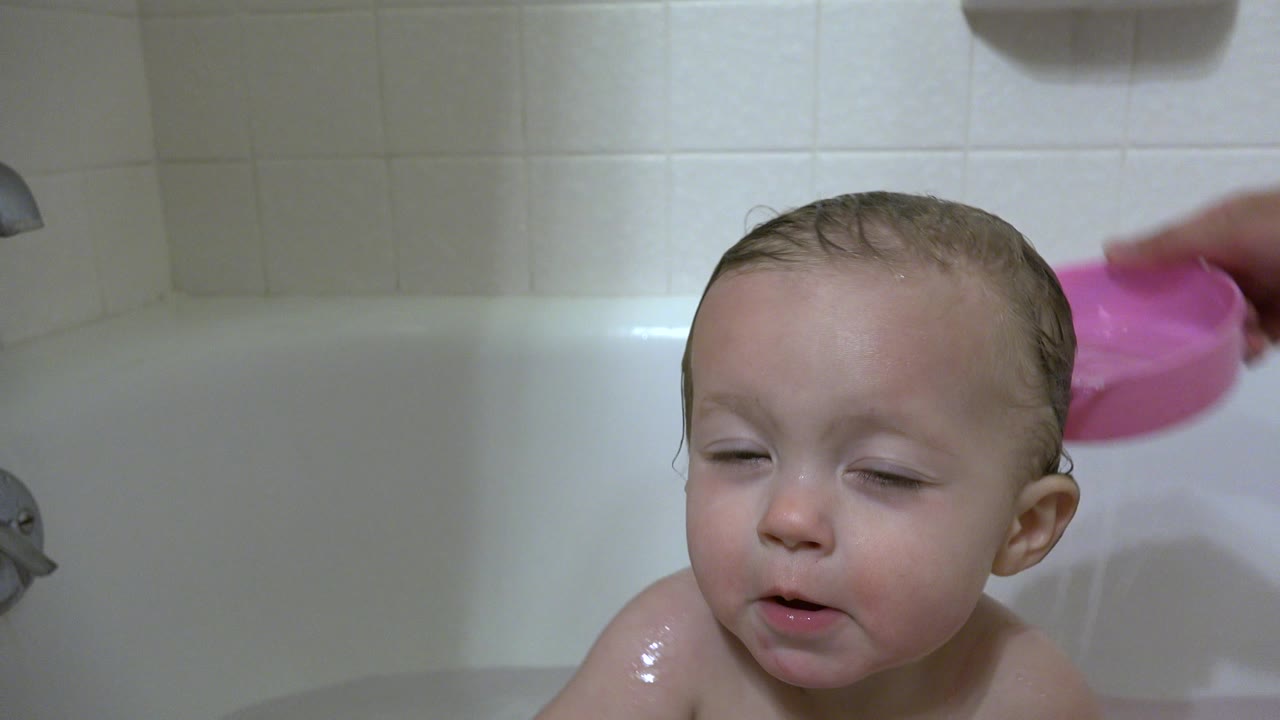 Wet baby in a bathtub as a pink bowl pours water nearby.