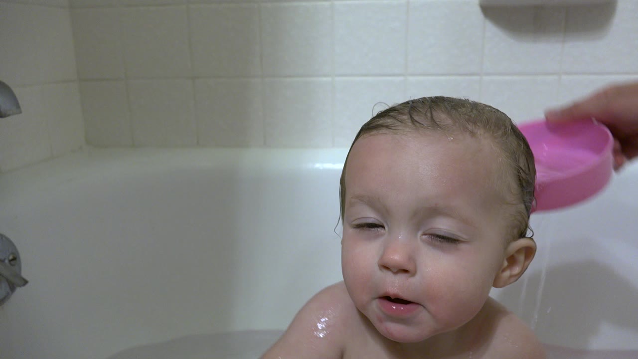 In Westminster, Colorado, the baby remains close to camera in the tub as a pink bowl tips above the shoulder area against pale bathroom tile.