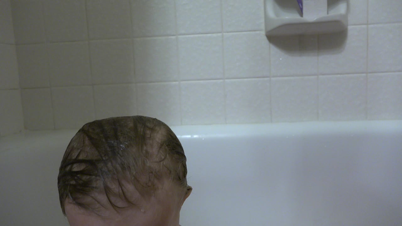 At the start of the Westminster, Colorado bath clip, only the wet top of a baby's head appears above the white tub edge beneath pale wall tile.