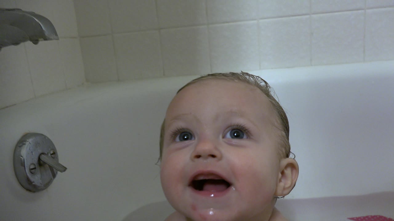 A wet baby in a white bathtub in Westminster, Colorado looks upward with an open mouth, with white tile and a metal faucet visible behind.