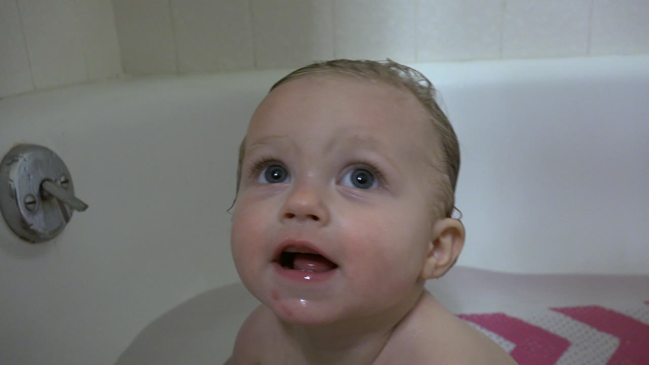 The baby shifts toward the right side of frame in the white bathtub, with wet hair, an open mouth, white tile, and part of a pink bath mat visible in Westminster, Colorado.