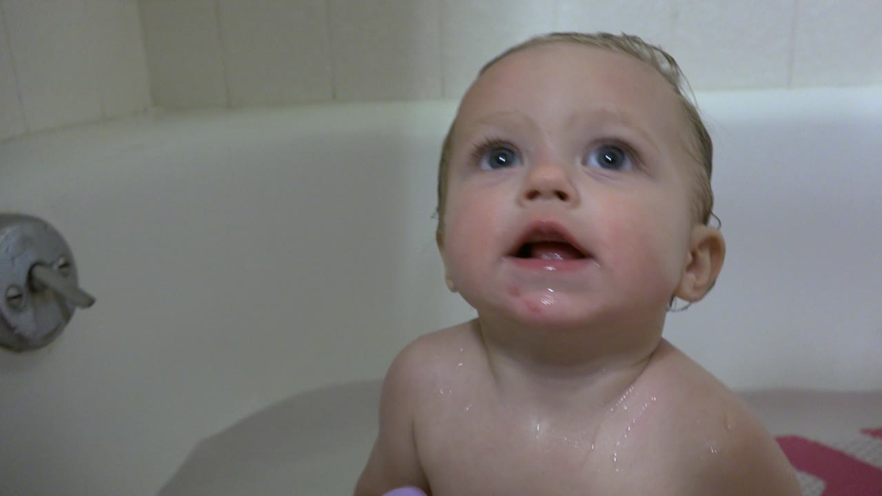 Baby sitting upright in bathtub water and looking above camera.