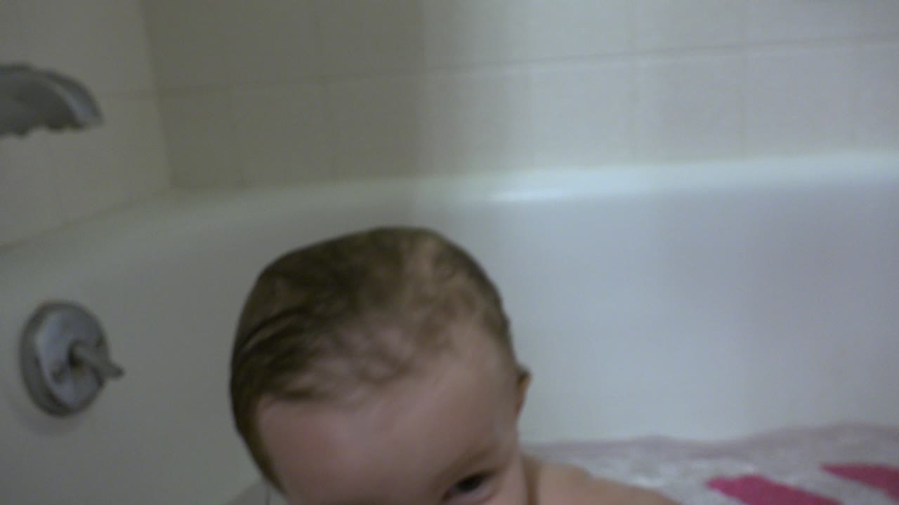 Wet infant low in a white bathtub beneath a metal faucet.