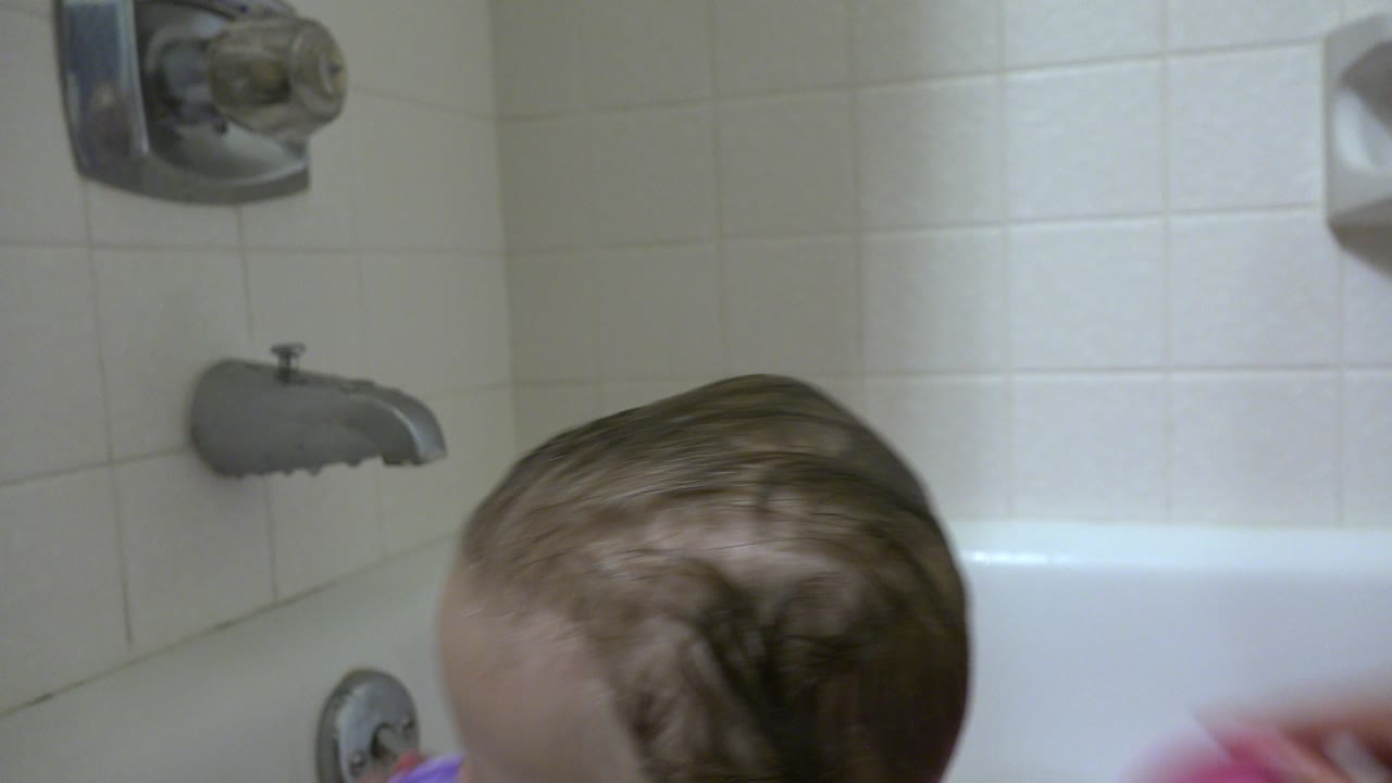 At 0:01 in Westminster, Colorado, the baby remains low in the bathtub while an adult hand brings a pink cup into the tiled bath scene.