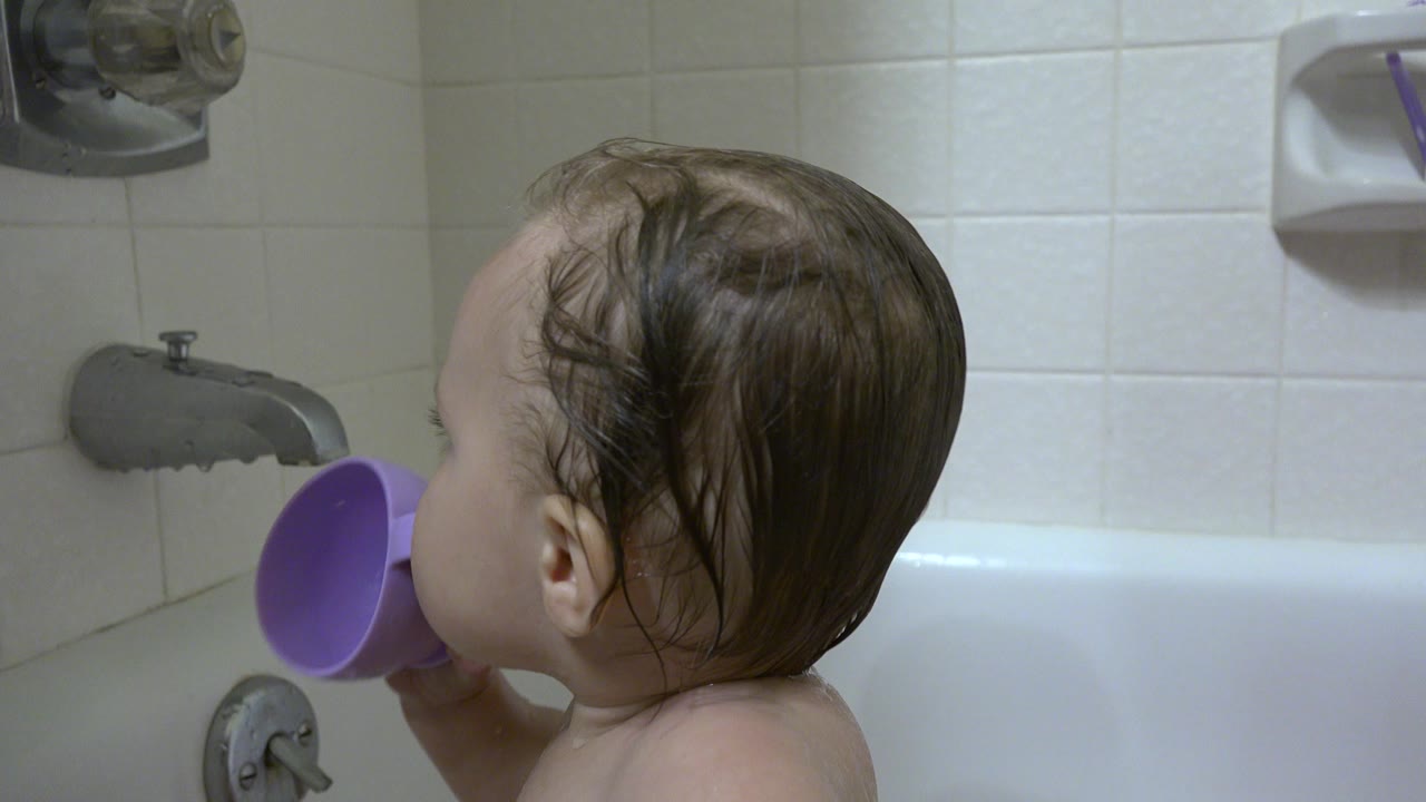 Toddler in a bathtub holding a purple cup near the tub spout.