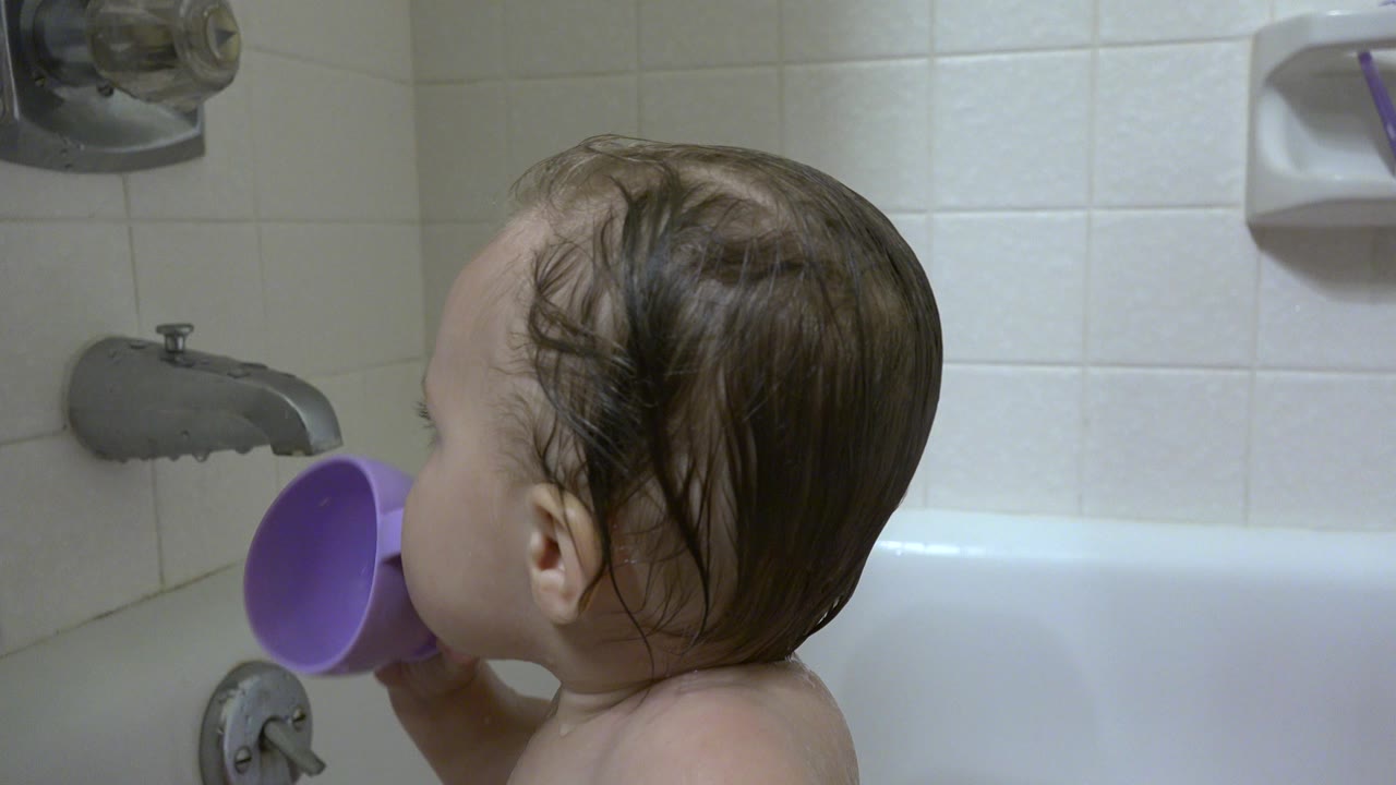 The toddler remains in profile in the Westminster bathroom tub, with wet hair, a purple cup, white tile, and metal fixtures visible.