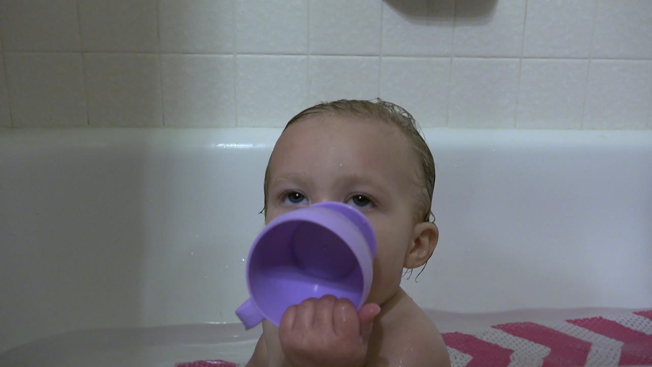 Baby boy in a bathtub holding a purple cup to his mouth.