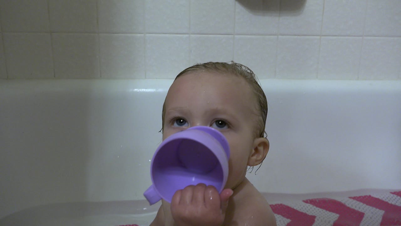 The baby boy remains centered in the bathtub in Westminster, Colorado, pressing the purple cup to his mouth with wet hair and tile behind him.
