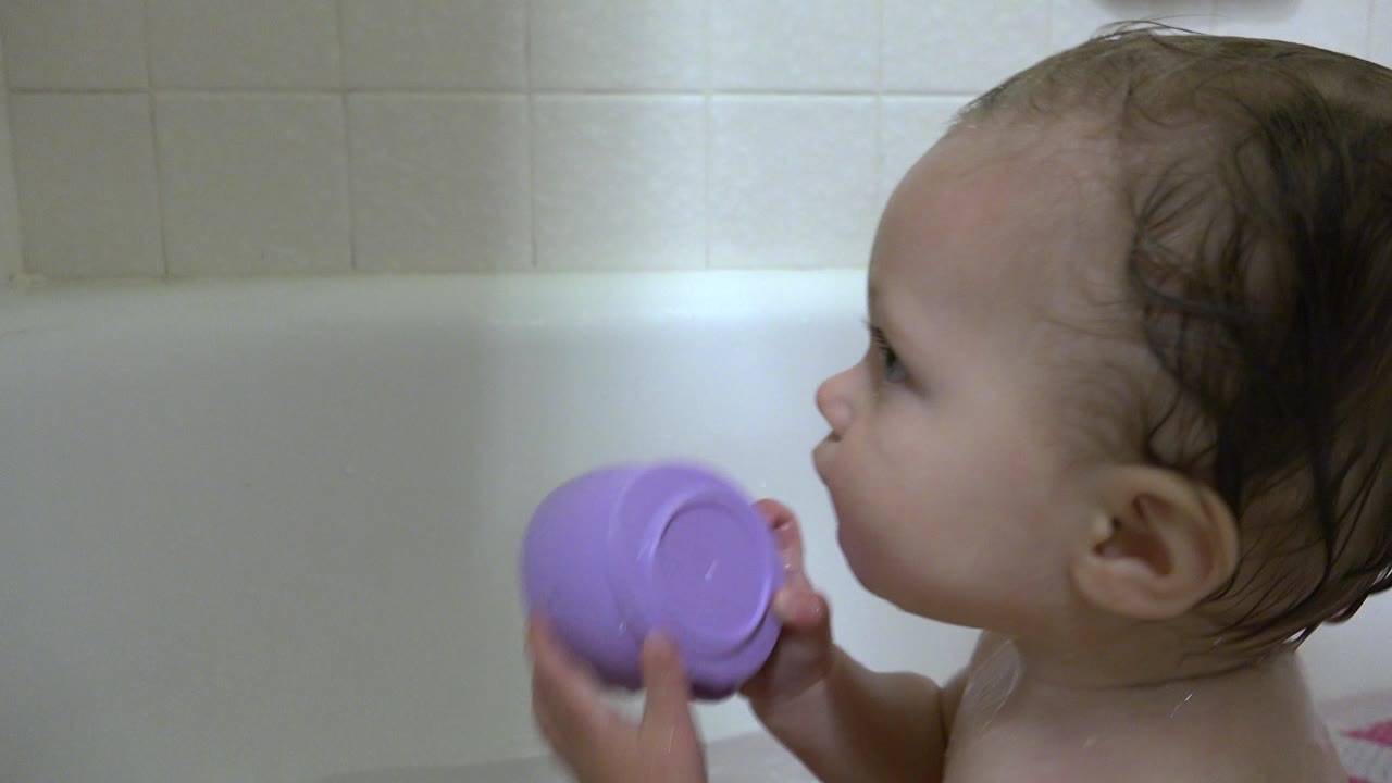 Wet-haired baby in a bathtub holding a violet plastic cup.