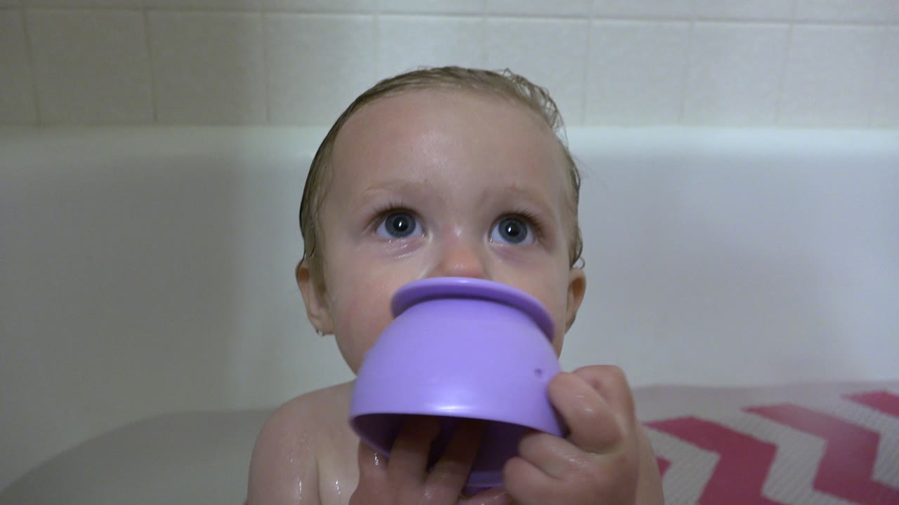 The baby remains centered in the Westminster bathroom tub, eyes lifted toward someone off camera while the purple toy covers part of the lower face.