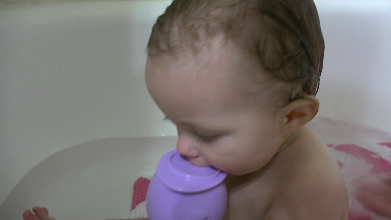 Baby in a bathtub holding a purple cup near the mouth.