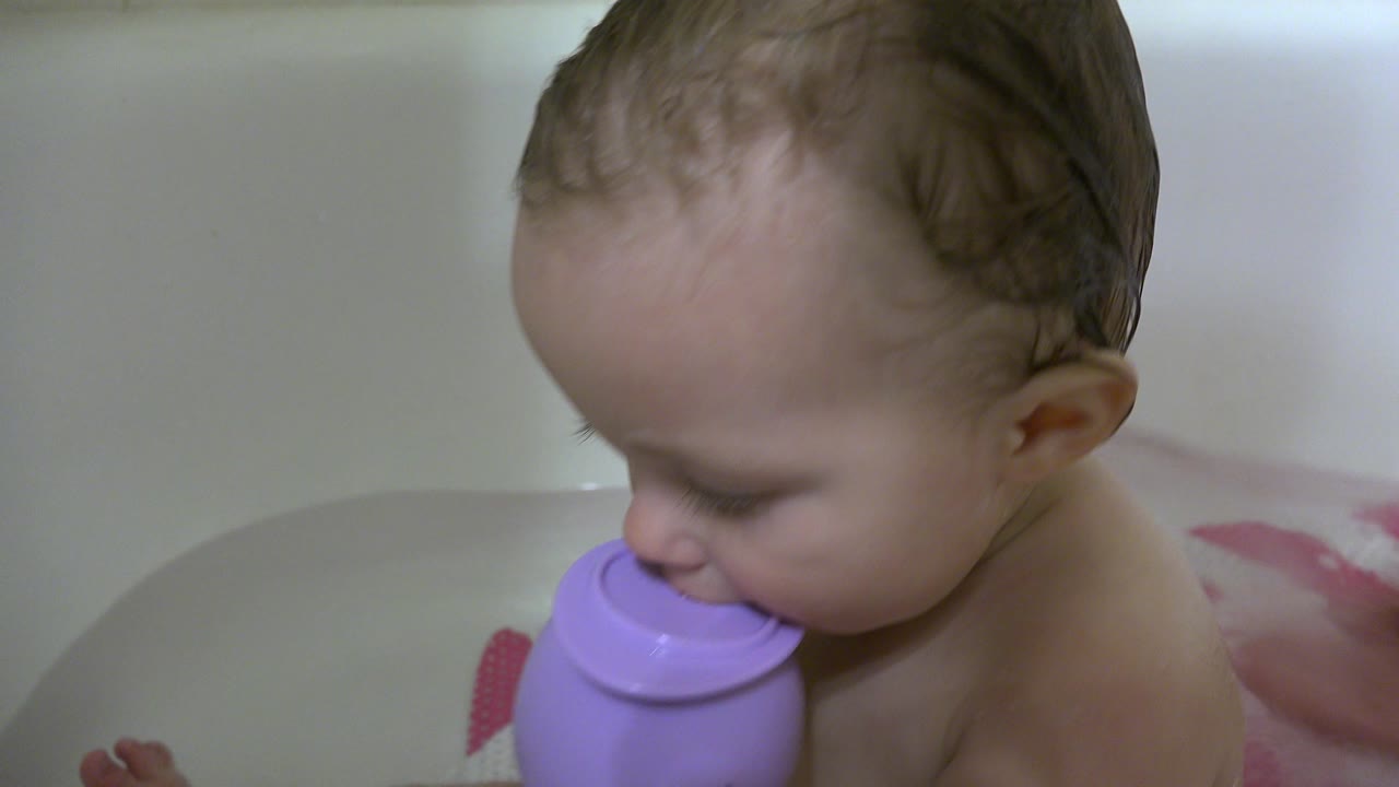 The baby remains close to the purple cup in the white bathtub, with soft bathroom light and pale tile behind the child in Westminster, Colorado.