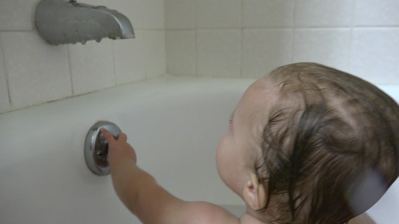 A wet-haired baby in a white bathtub in Westminster, Colorado reaches toward a chrome bath handle below a tub spout.