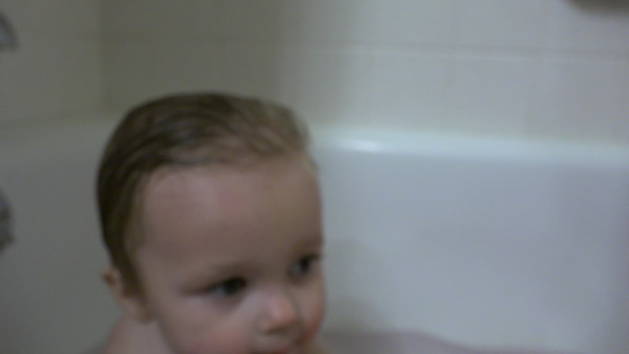 At 0:01, the toddler remains in the white bathtub in Westminster, Colorado, looking off to the side beneath soft indoor bathroom light.