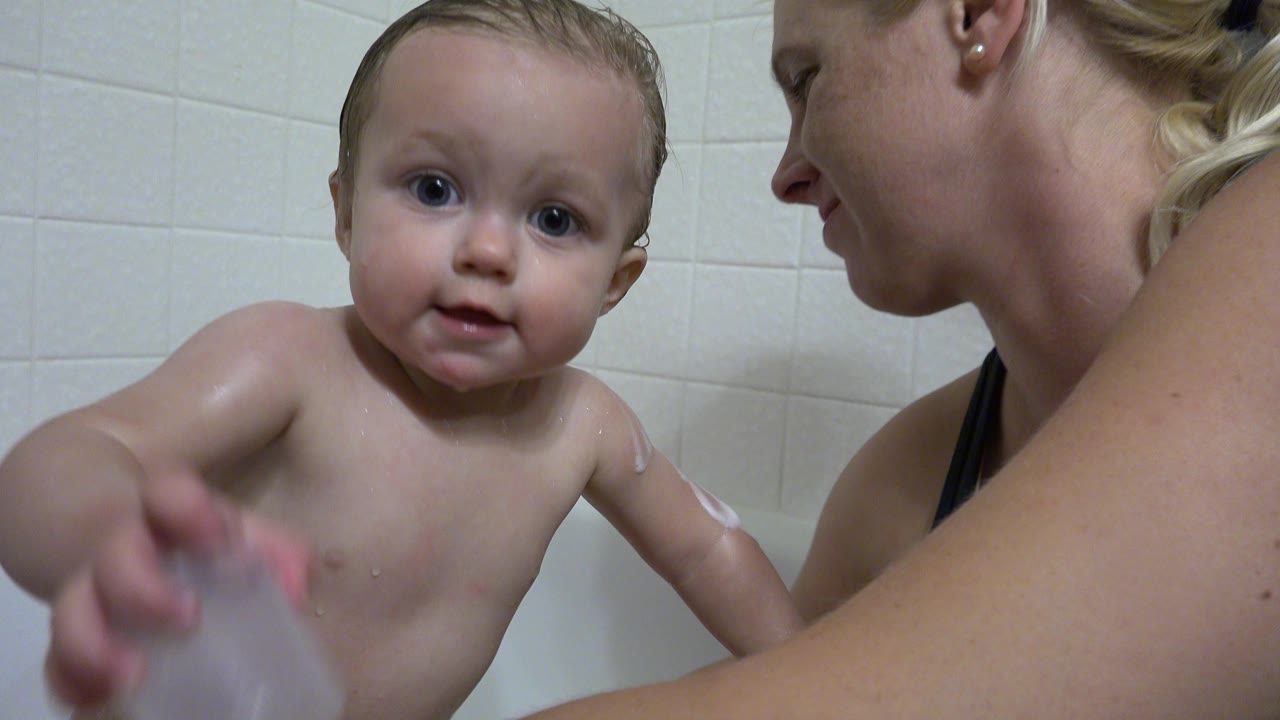 In Westminster, Colorado, a wet baby sits close to the camera in a white tiled bathtub while an adult woman is cropped at the right edge.