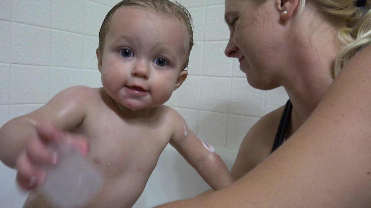 In Westminster, Colorado, the baby looks toward the camera and reaches forward with a translucent bath cup as the mother watches from the side.