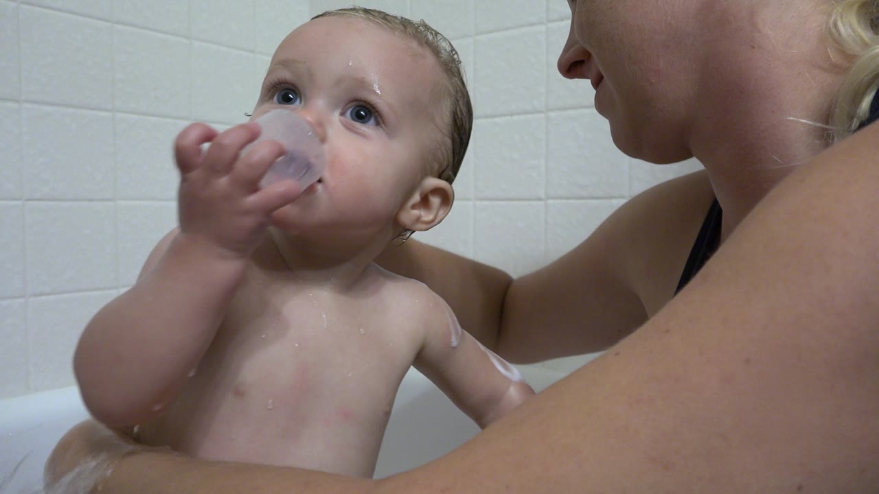 Baby puts a translucent cup to their mouth in the bathtub.
