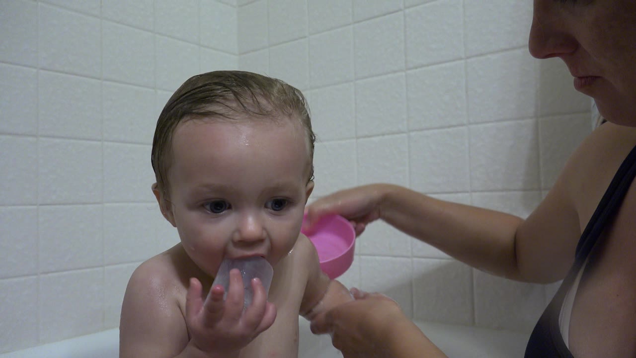 Young child in bathtub holding a gray cup while caretaker washes nearby.
