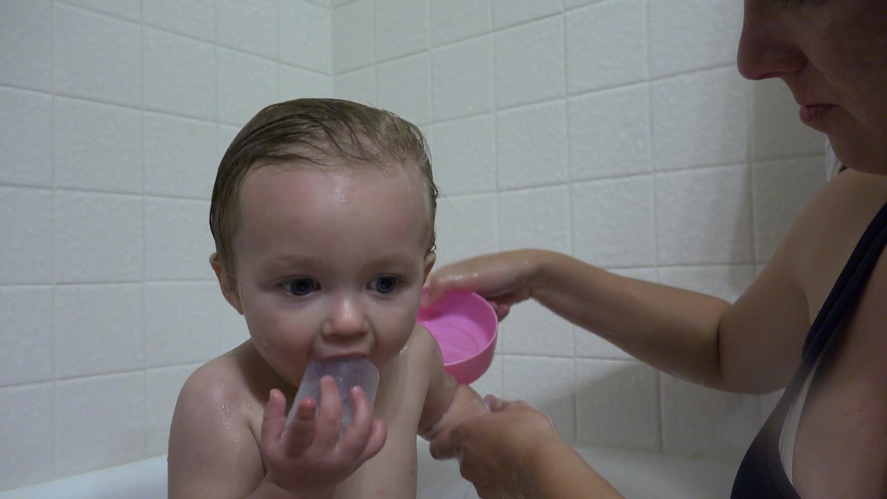 At 0:01 in Westminster, Colorado, the child looks toward the camera with a gray cup at their mouth while the adult caretaker holds a pink cup near the tub wall.