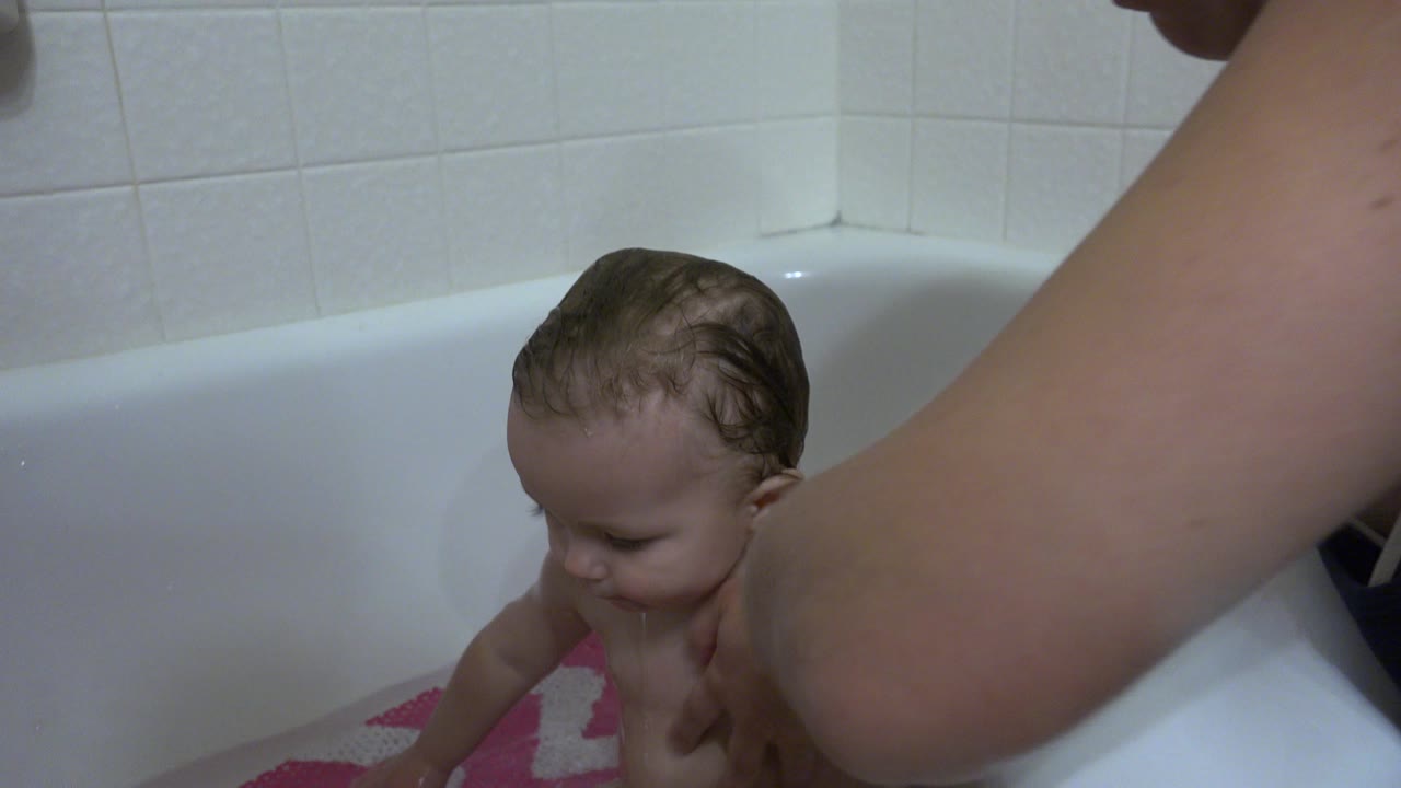 At 0:00 in Westminster, Colorado, a wet baby sits low in a white bathtub while an adult caregiver's arm crosses the foreground near the tub wall.
